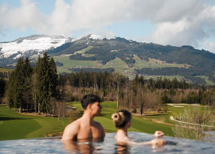 Pärchen schwimmt im Infinitypool mit Blick auf den Golfplatz und Berge