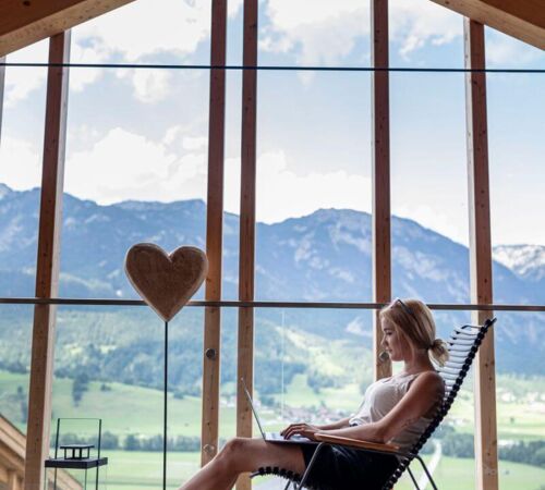 Eine junge Frau sitzt im Schaukelstuhl in ihrem Ferienhaus und arbeitet am Laptop.