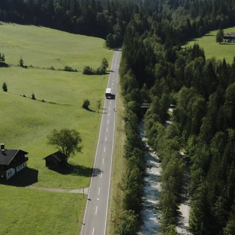 Bus fährt auf einer Landstraße umgeben von grünen Wiesen, Wald und Fluss.