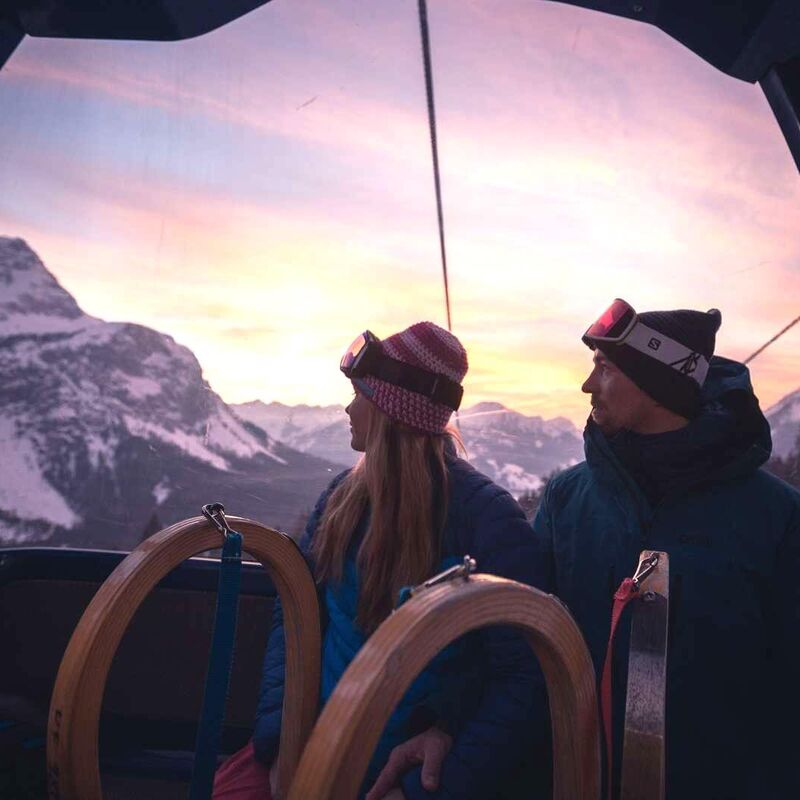 Ein Pärchen mit 2 Schlitten blickt aus der Gondelbahn auf die Berge der Tiroler Zugspitz Arena im Sonnenuntergang.