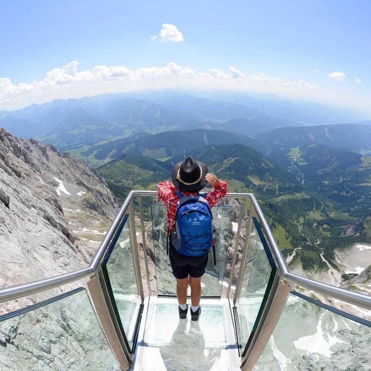Ein Wanderer steht auf einer Glasplattform am Dachstein und genießt die atemberaubende Aussicht ins Tal.