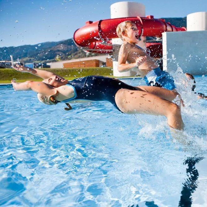 Children joyfully splash in a pool while playing near a water slide in a sunny outdoor setting.