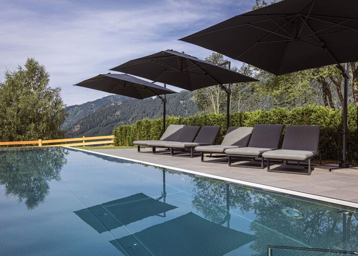 Moderner Infinitypool mit extrabreiten Sonnenliegen, weich gepolsterten Auflagen und großen Sonnenschirmen sowie traumhaftem Ausblick auf die grüne Berglandschaft im Salzburger Land.