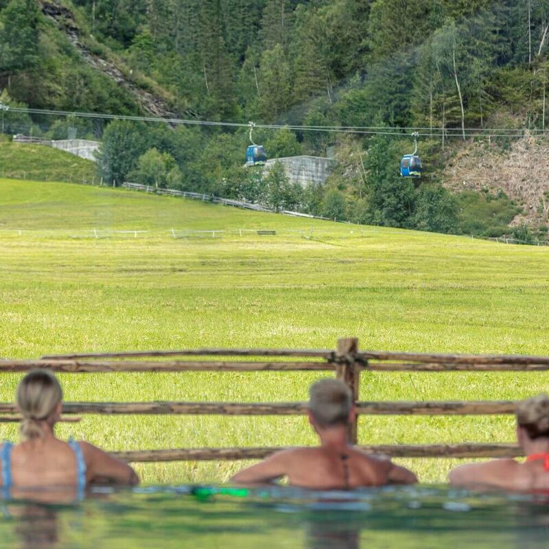 3 Freunde genießen den Blick auf die Bergbahn vom Pool aus.