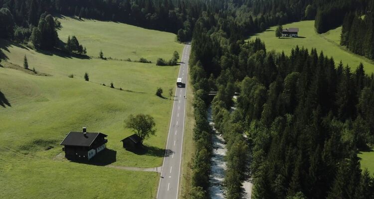 Bus travelling on a country road surrounded by green meadows, forest and river.