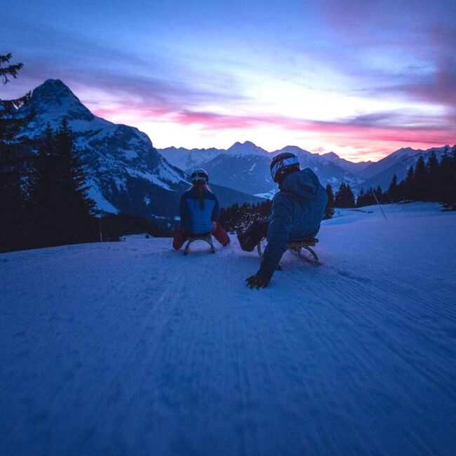 2 tobogganists ride their sledges down the toboggan run at dusk.