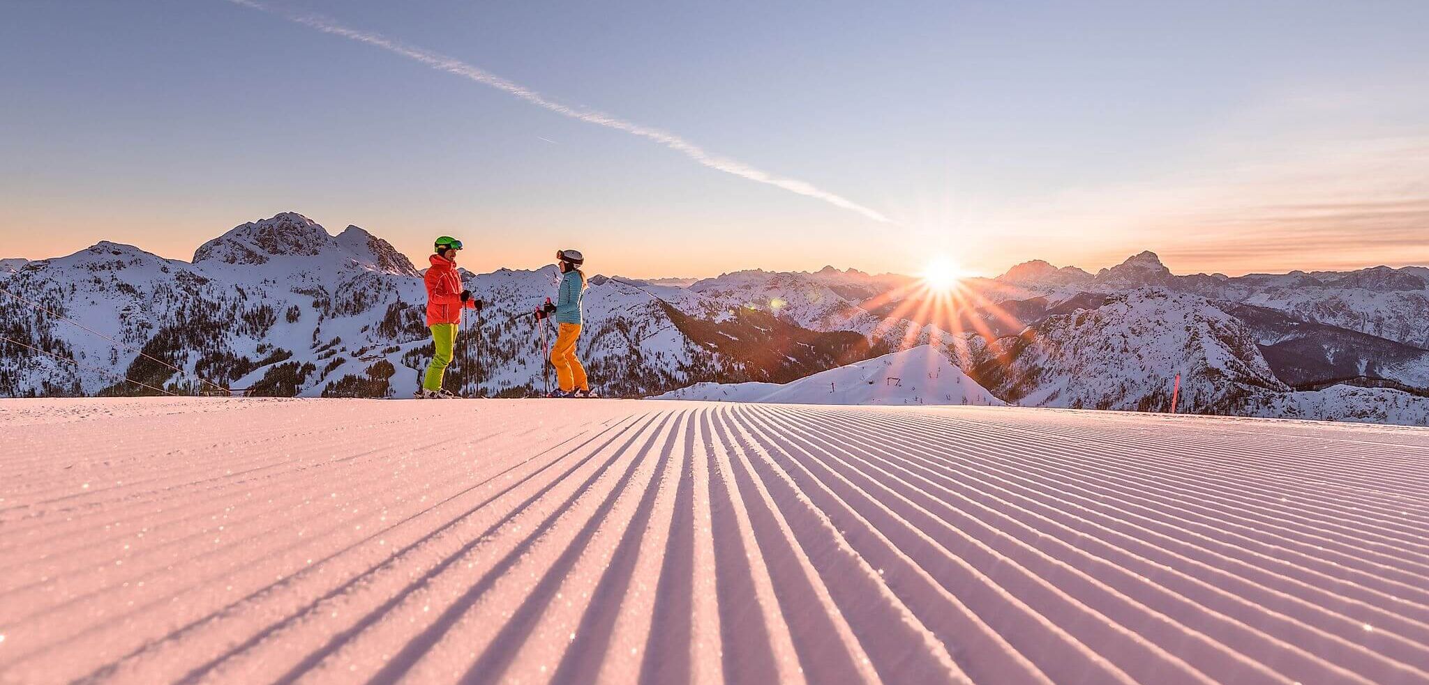 Zwei Skifahrer auf einer perfekt präparierten Piste im Skigebiet Nassfeld/Kärnten, Bergpanorama und Sonnenaufgang im Hintergrund.