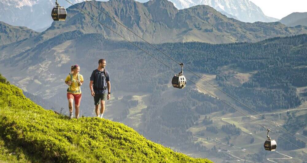 The summer mountain railway runs above 2 hikers, with imposing mountain ridges in the background.
