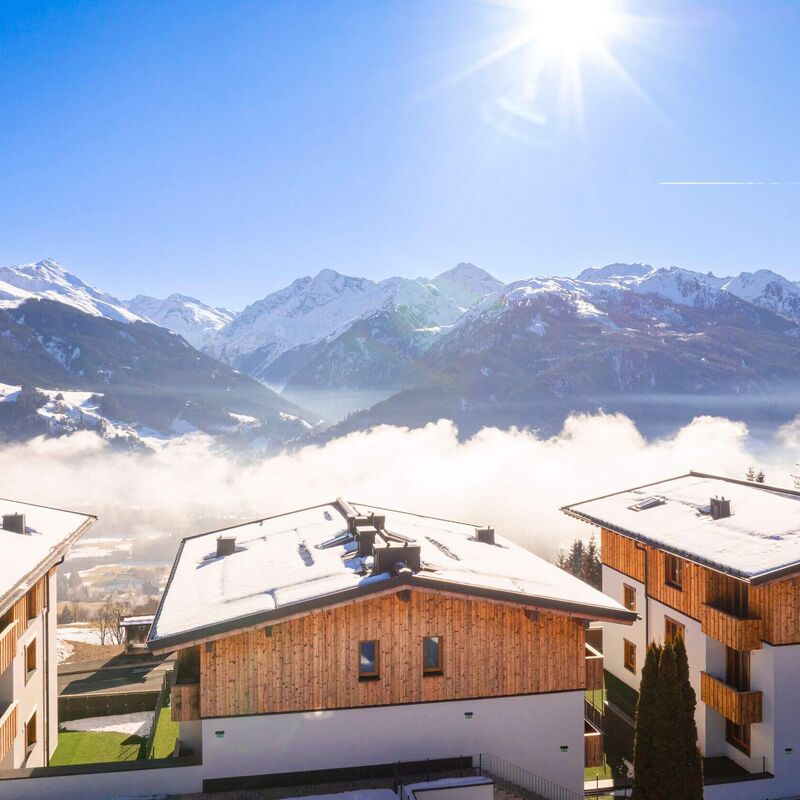 Snow covered buildings overlook a breathtaking valley and majestic mountains under a clear blue sky.