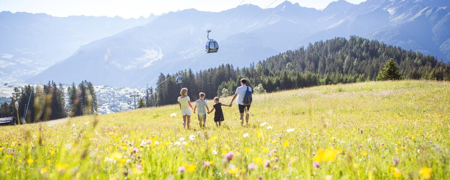 Eine Familie wandert über eine Bergwiese, oberhalb fährt eine Gondelbahn und bietet einfachen Zugang zum Wandergebiet