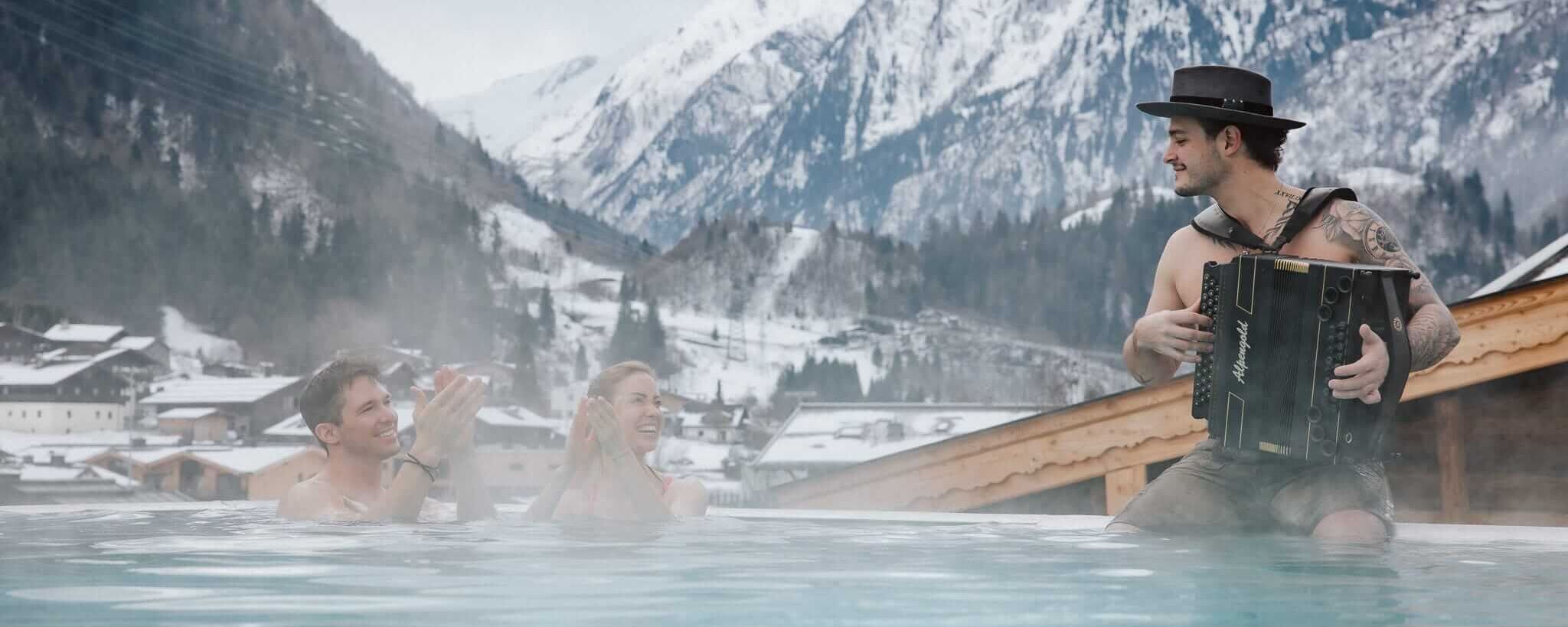 Mensen genieten van een warmwaterbron met livemuziek en bergen op de achtergrond in winterse sneeuw. | © Markus Laubvogel