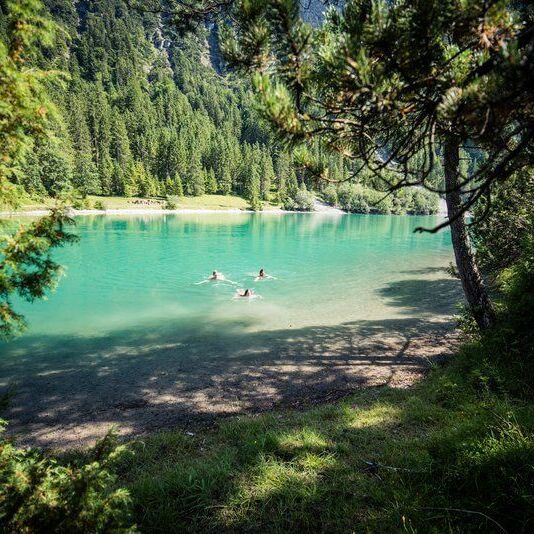 Three people swimming in a turquoise mountain lake surrounded by forest