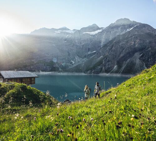 Zwei Wanderer spazieren an einer urigen Berghütte vorbei, entlang einer blühenden Almwiese mit Blick auf einen türkisfarbenen Stausee und die umliegende Bergkulisse im Salzburger Land.