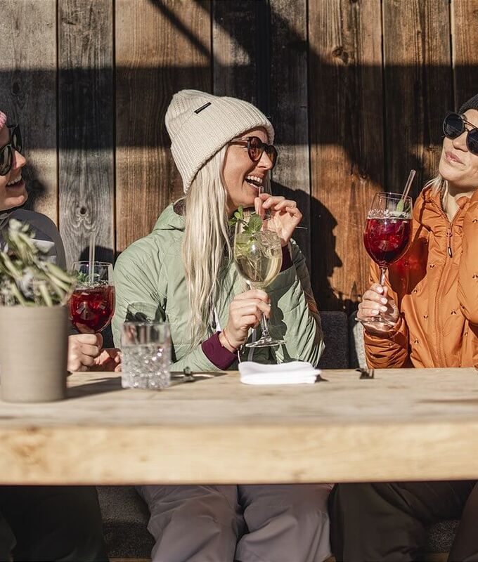 3 Freundinnen im Ski Outfit sitzen vor einer Skihütte in der Sonne.