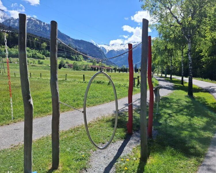 A circular play feature stands amidst scenic mountains and green fields along the Motorikweg in Kaprun.