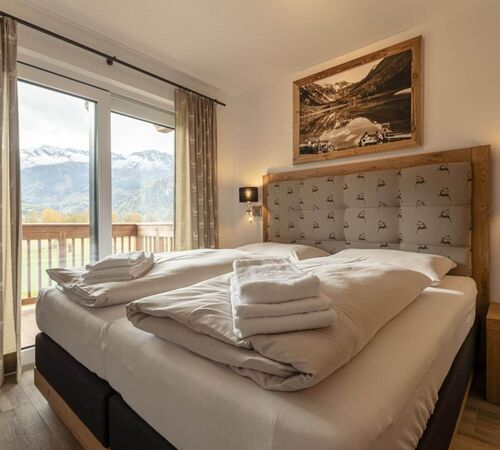 Helles Schlafzimmer mit bequemen Boxspring Doppelbett, Holzelementen, gefalteten Handtüchern auf dem Bett, gepolstertem Kopfteil, Nachttischen und Balkon mit Aussicht auf herbstliche Natur und verschneite Berge