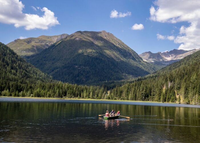 Grüner Bergsee mit Ruderboot umgeben von Wäldern mit Berg im Hintergrund