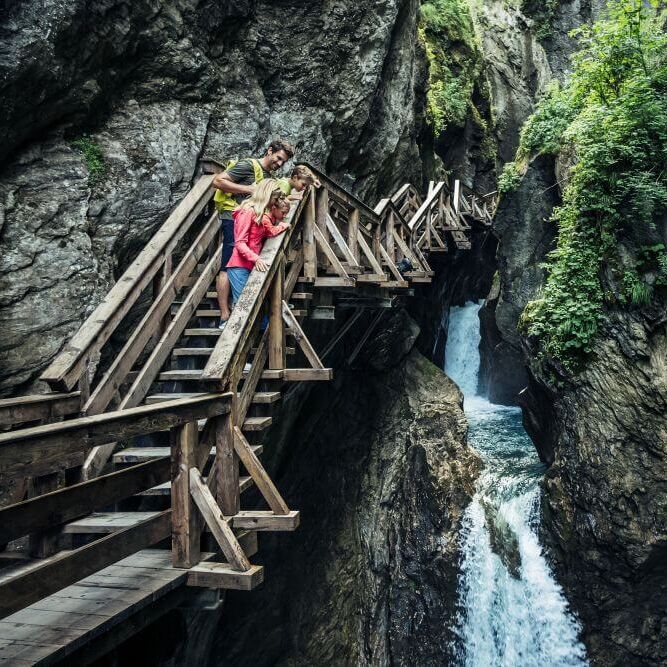 A family admires a waterfall from a wooden bridge in the scenic Sigmund Thun Klamm gorge.