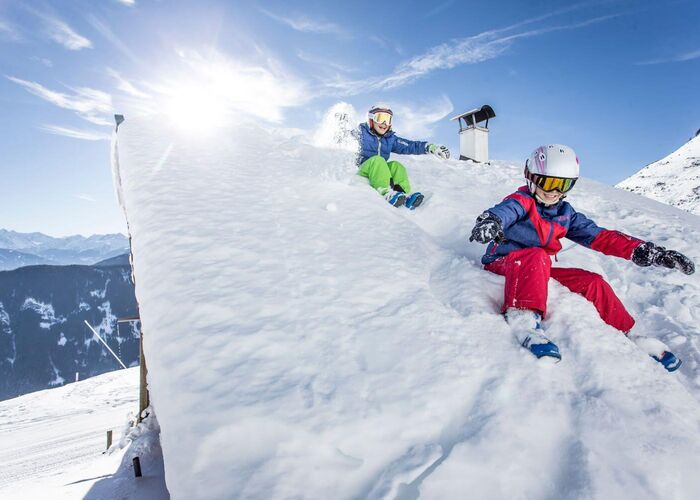 Children in ski outfits slide off the snow covered roof of an alpine hut.