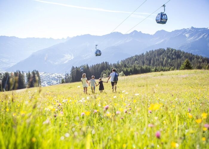 Familie wandert auf einer Bergwiese, darüber schwebt die Gondelbahn.