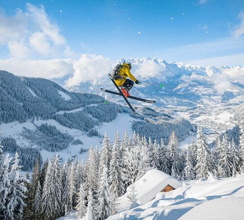 Ein Skifahrer springt durch glitzernden Pulverschnee vor der Kulisse verschneiter Berge, Täler und Wälder im Salzburger Land – Winterfreude pur in unberührter Natur.