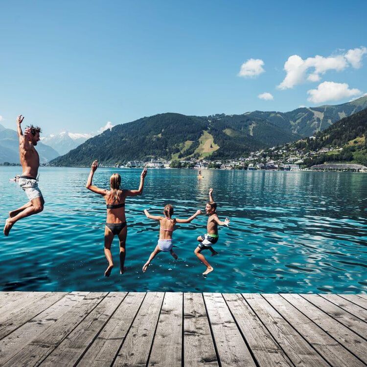 Eine Familie springt fröhlich in den Zeller See an einem sonnigen Tag mit Bergen im Hintergrund. | © Zask