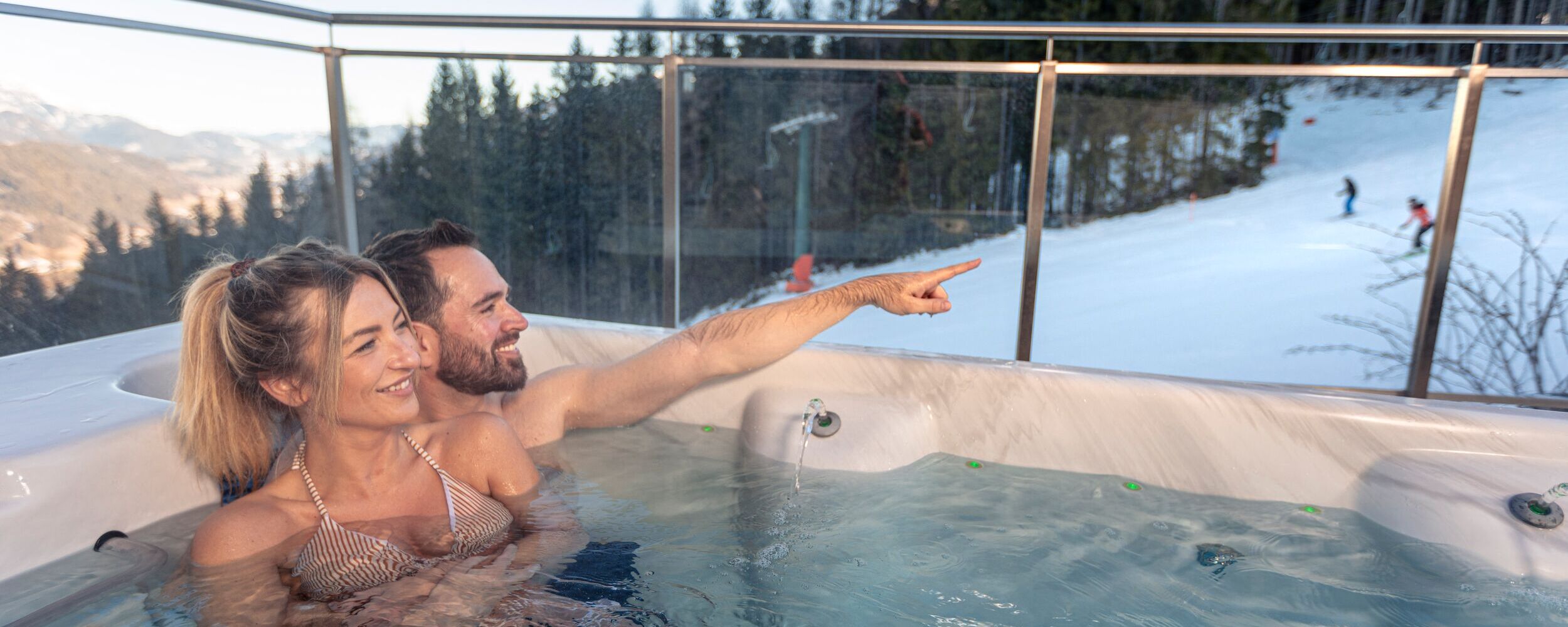 Ein Paar entspannt im Whirlpool und blickt auf Skifahrer im Schnee mit Bergen im Hintergrund.