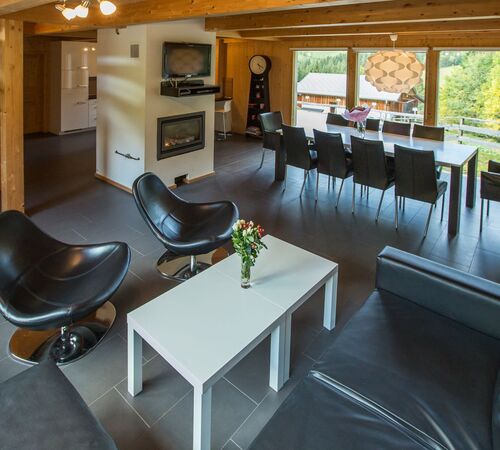 Großzügiger Wohnraum im Ferienhaus mit Kamin, Ledersesseln und Ledercouch, Essbereich mit Tisch und Stühlen, großen Panoramafenstern mit Blick auf die umliegende Berglandschaft
