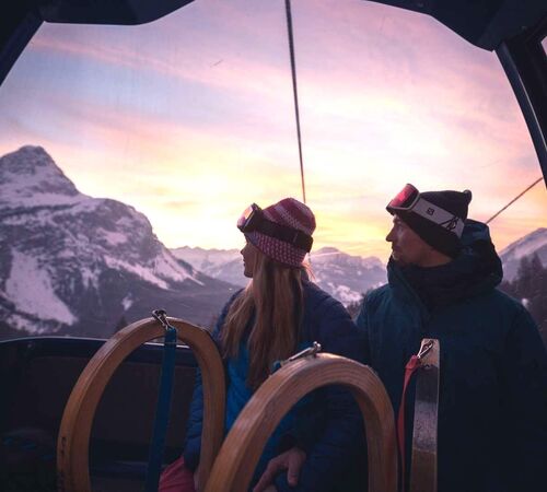 A couple with 2 sledges look out of the gondola lift at the mountains at sunset.