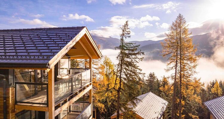 Moderne Holzchalets über den Wolken, umgeben von herbstlichen Bäumen und Bergen im sonnigen Licht.