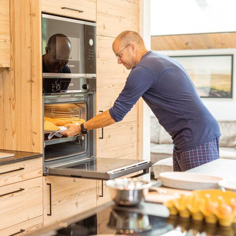 Mann holt frisch aufgebackene Brötchen für das Frühstück aus dem Backofen.