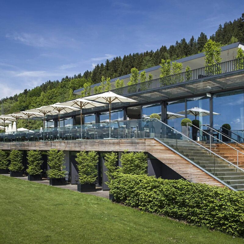 Moderne Restaurantansicht mit Glasfassade und sonnigen Terrassen in einer grünen Umgebung.