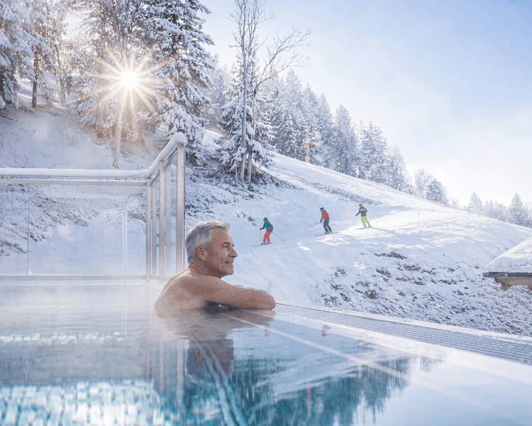 Ein Mann entspannt im Thermalwasser, während Skifahrer auf einer verschneiten Piste vorbeifahren.