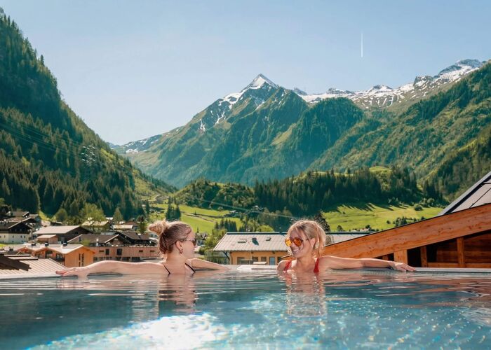 Twee vrouwen ontspannen in een zwembad met uitzicht op bergen en een helderblauwe lucht.