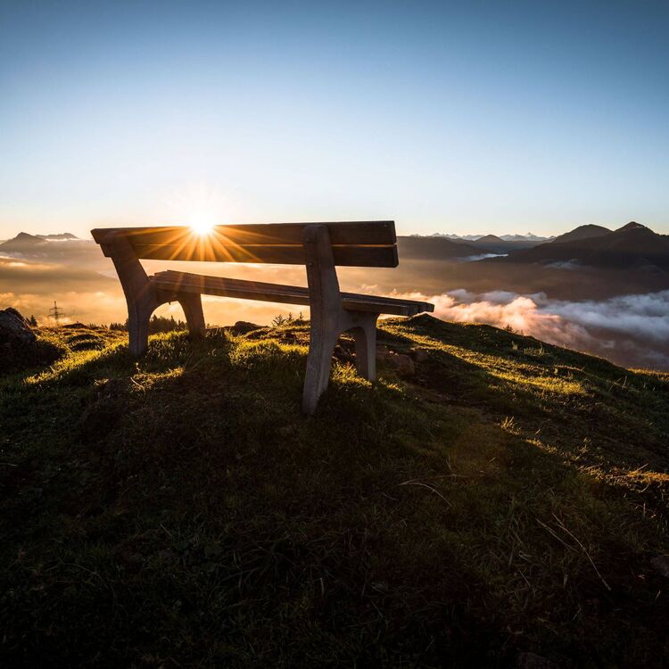 Eine Holzbank auf einem Tiroler Gipfel bei Sonnenaufgang über den Bergen und Wolken.