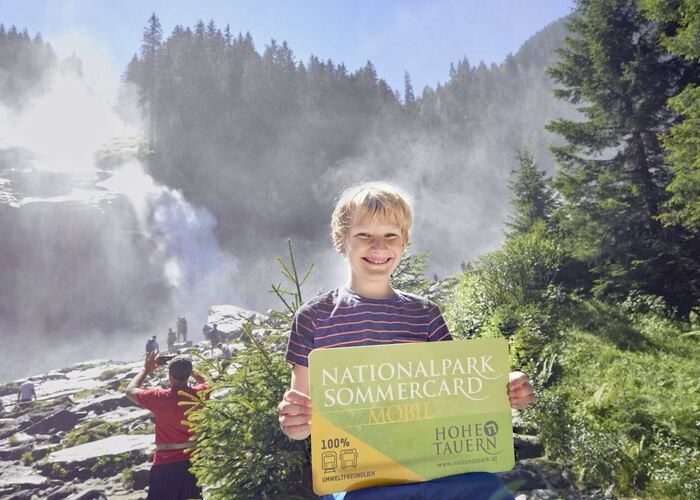 Junge steht mit der Nationalpark Sommercard in der Hand vor den Krimmler Wasserfällen.