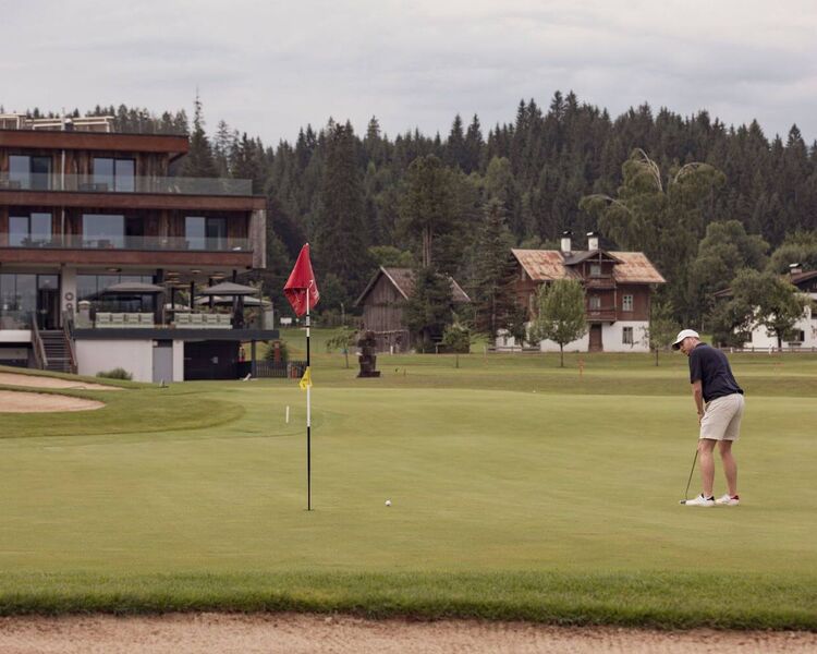 A golfer putts on a lush green course at Westendorf, with mountains and a modern clubhouse in the background.