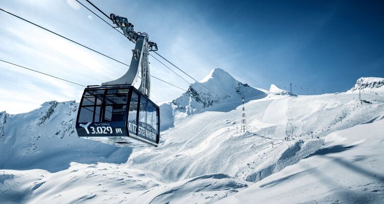 Die Kitzsteinhorn-Gipfelbahn schwebt über verschneite Hänge bei strahlendem Himmel und majestätischen Bergen.