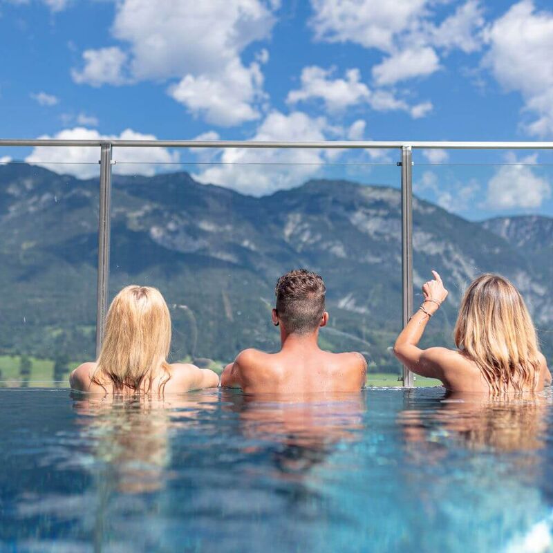 3 Freunde schwimmen im Pool und blicken auf das Dachstein Massiv