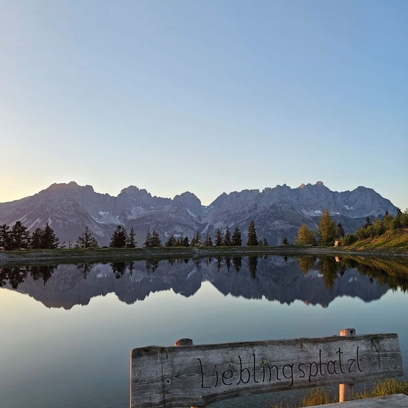 The Wilder Kaiser is reflected in the Astbergsee lake at sunset, with a wooden bench in the foreground.