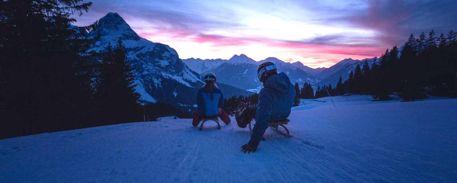 2 tobogganists ride their sledges down the toboggan run at dusk.