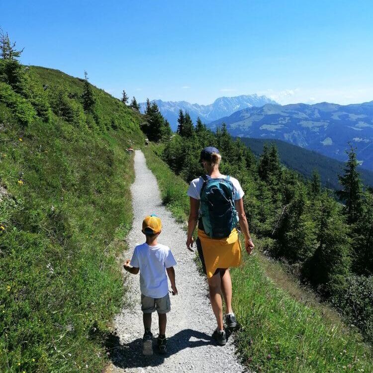 Eine Frau und ein Junge wandern einen schmalen Weg durch grüne Hügel unter klarem Himmel.