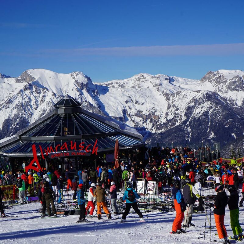 Skifahrer und Besucher versammeln sich vor der Almrausch Hütte in Planai, umgeben von schneebedeckten Bergen.
