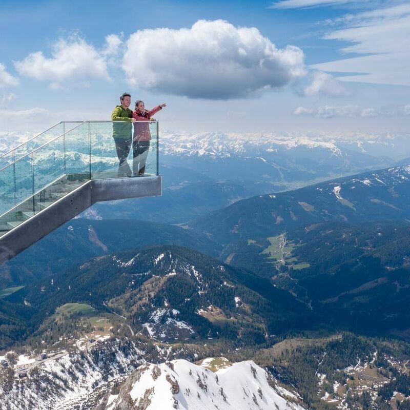 Two people stand on the Dachstein Himmelsleiter viewing platform, admiring stunning mountain scenery.