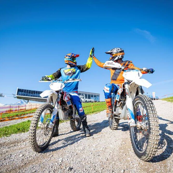 Two motocross riders celebrate with a high five on a sunny day at Schmittenhöhe, surrounded by scenic mountains.