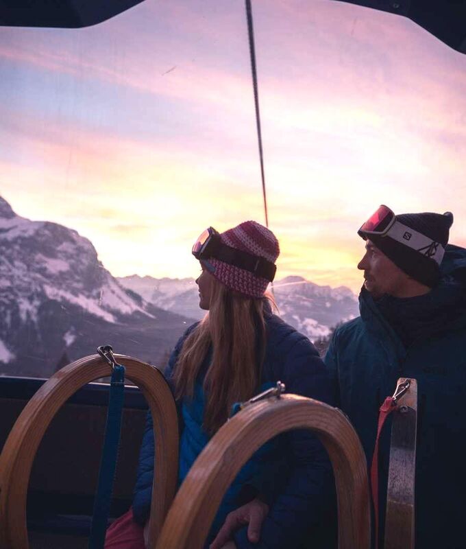 Ein Pärchen mit 2 Schlitten blickt aus der Gondelbahn auf die Berge der Tiroler Zugspitz Arena im Sonnenuntergang.