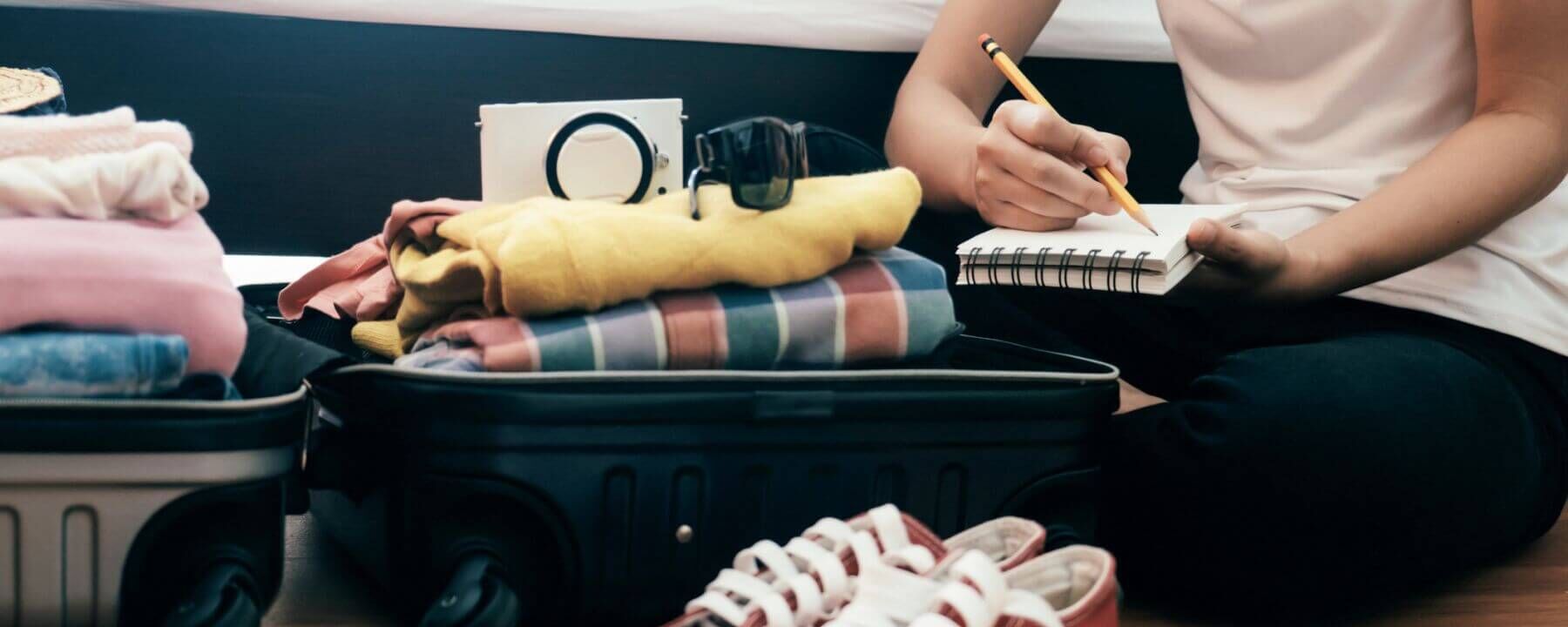 Junge Frau mit einer Checkliste sitzt vor ihrem offenen, vollbepackten Koffer auf dem Boden.