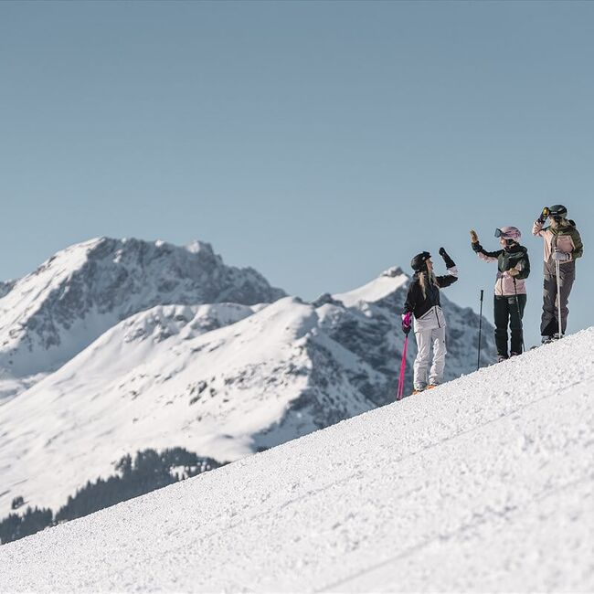 3 skiers stand on the firn snow piste and clap their hands.