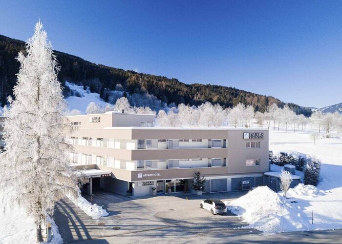 Winter driveway with parked cars in front of the Tauernresidence Radstadt, with a hoar frosted tree and snowy meadows next to it