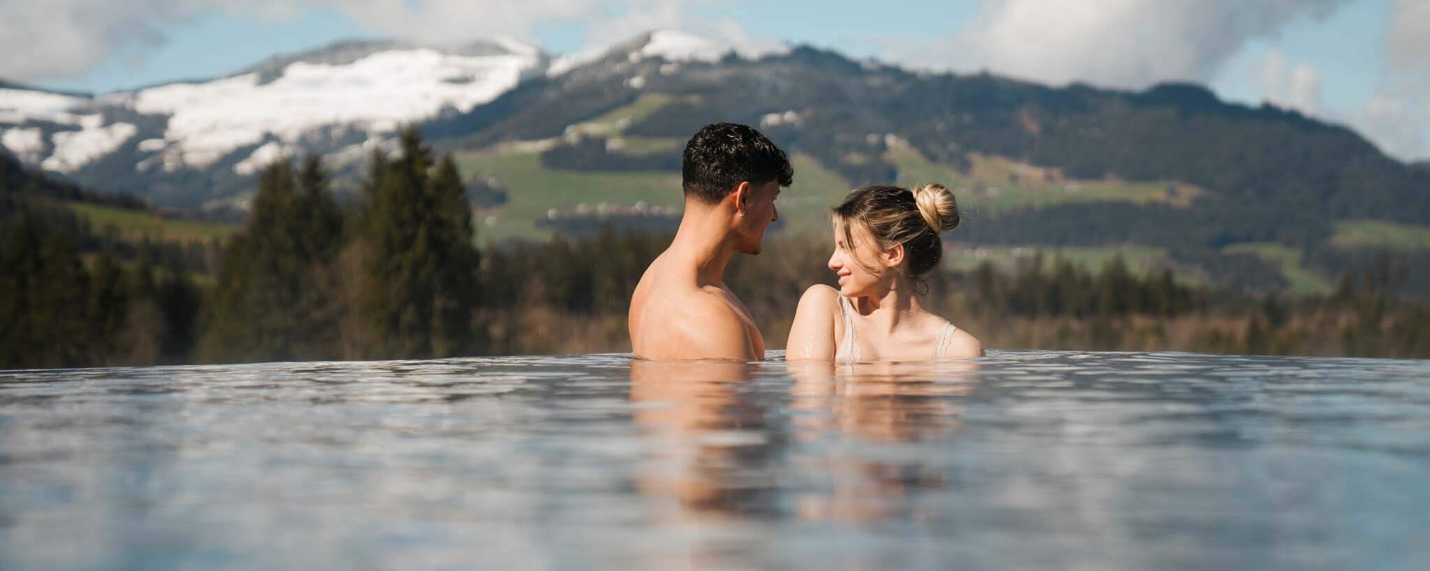 Een stel ontspant samen in een buitenzwembad met uitzicht op de Alpen.